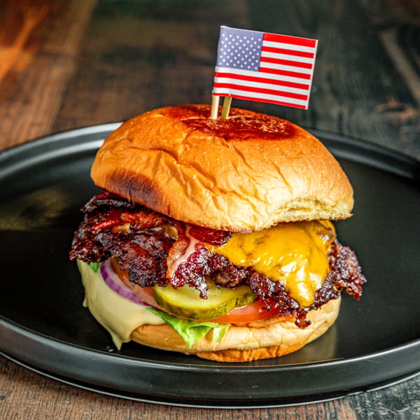 A juicy International Burgers bacon cheeseburger with an American flag toothpick on a dark plate.