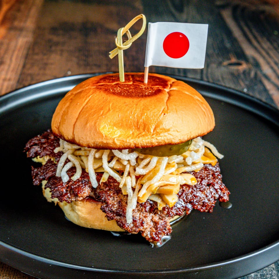 A Japanese-themed burger with a flag, crispy onions, and pickles on a black plate.