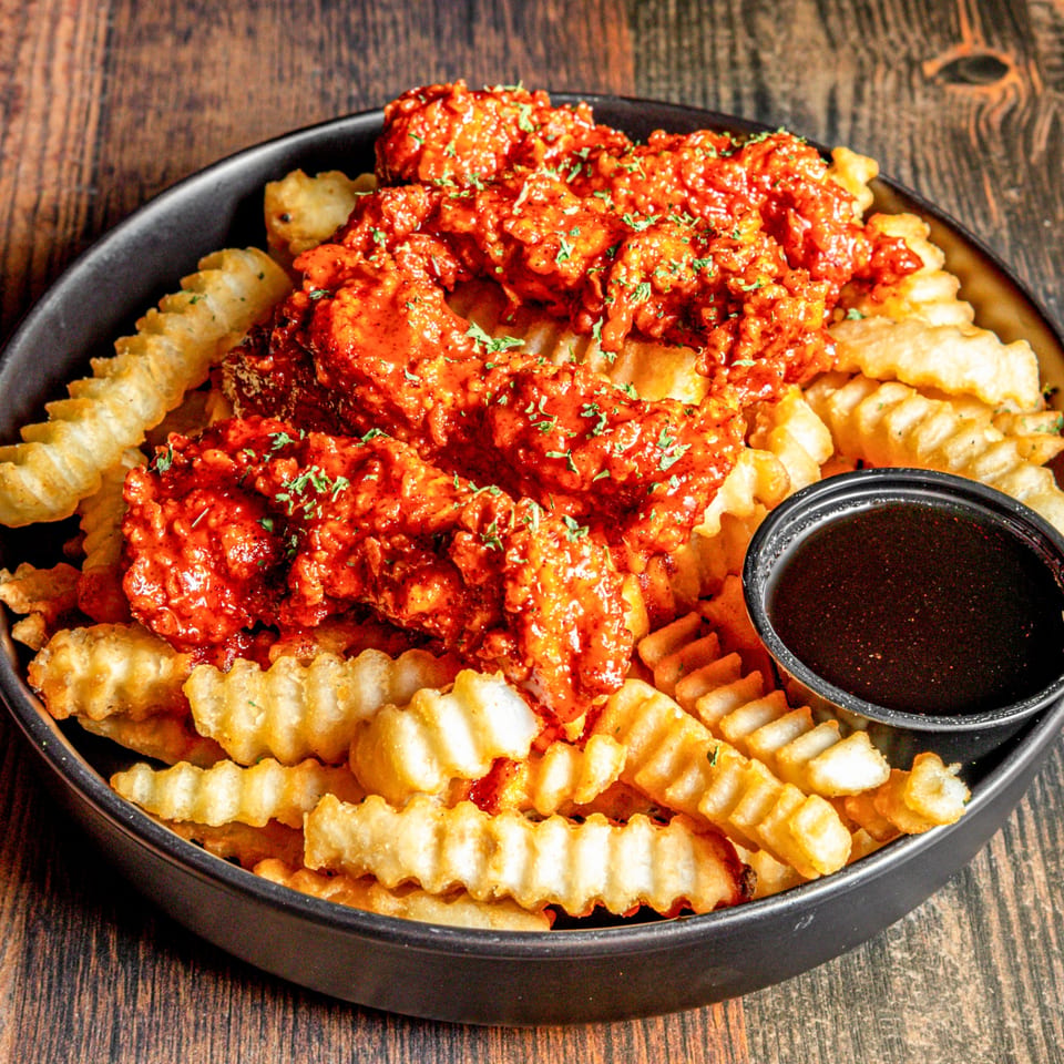 Spicy chicken tenders with crinkle-cut fries and a side of dark dipping sauce in a black bowl.