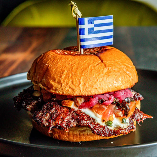 A Greek-themed burger with a flag, pickled onions, and melted cheese on a dark plate.