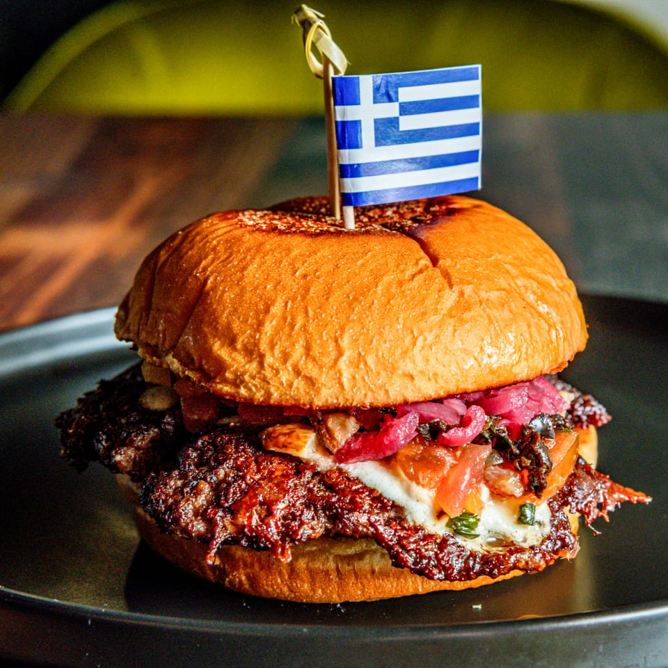 A Greek-themed burger with a flag, pickled onions, and melted cheese on a dark plate.