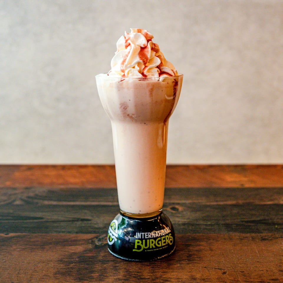 Strawberry milkshake with whipped cream and syrup drizzle, in an International Burgers branded glass.