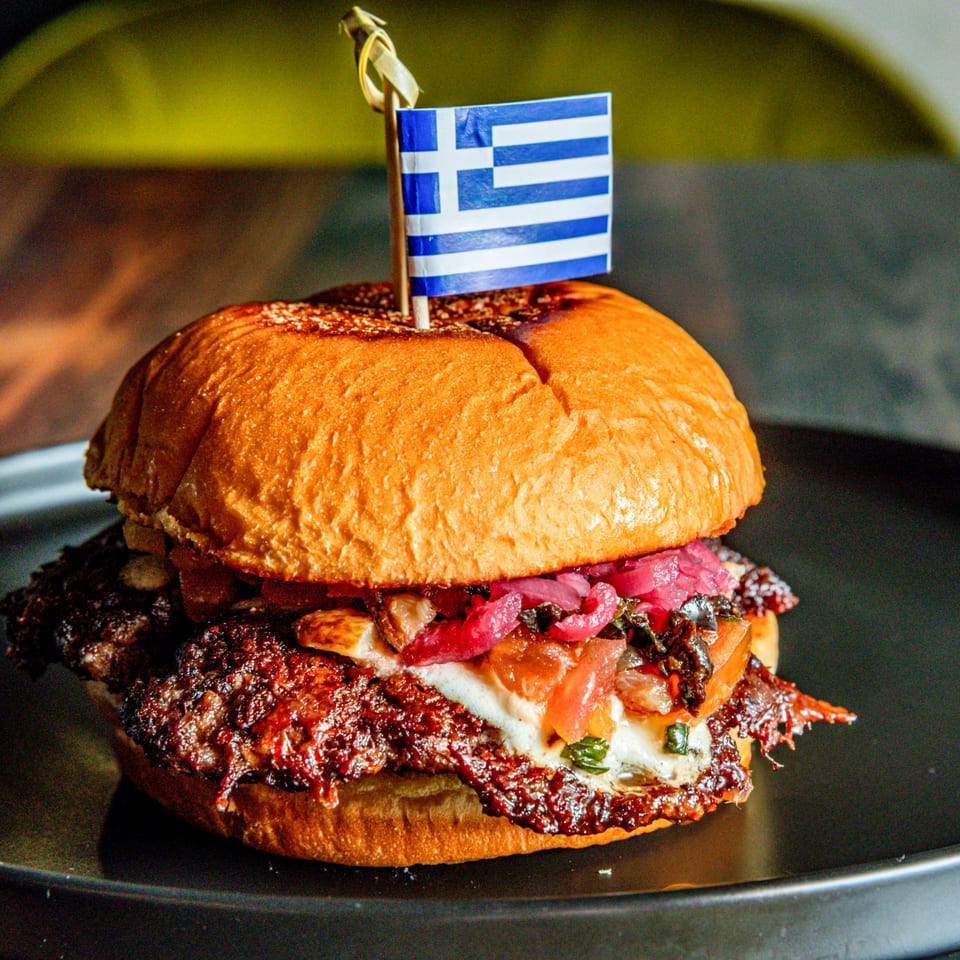 A Greek-themed burger with a small Greek flag on top, featuring a patty, red onions, and sauce.