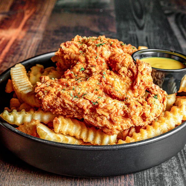 Crispy fried chicken tenders and crinkle-cut fries in a black bowl with a side of dipping sauce.