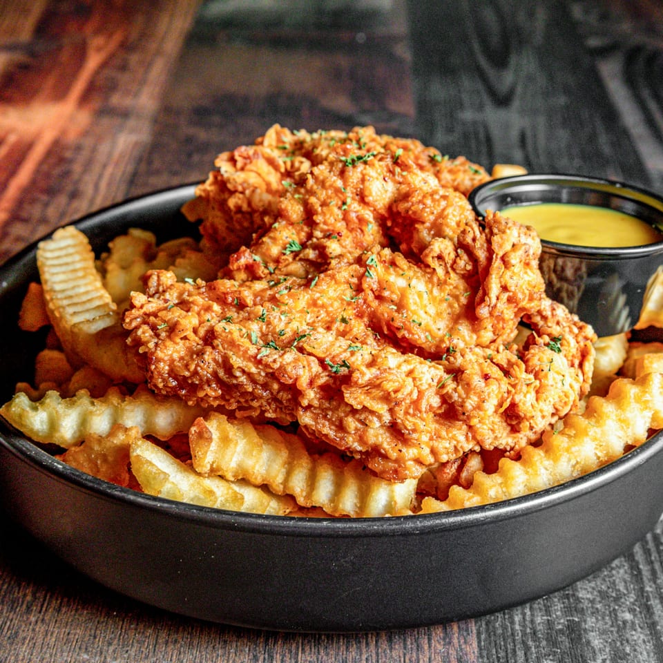 Crispy fried chicken tenders and crinkle-cut fries in a black bowl with a side of dipping sauce.
