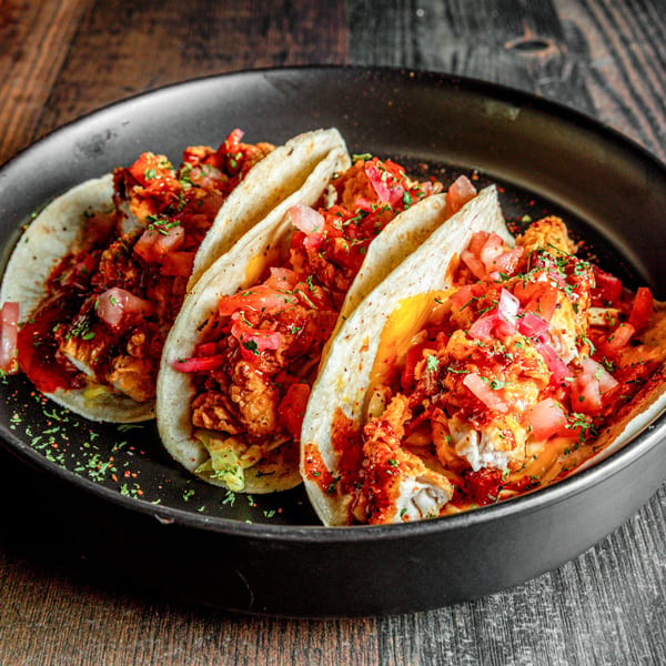 Three vibrant tacos filled with crispy chicken, red sauce, diced tomatoes, and herbs in a black pan.