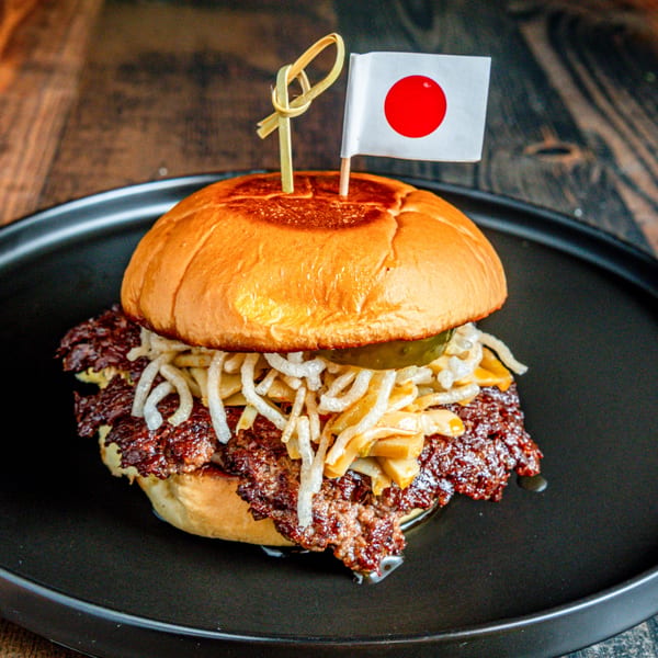A Japanese-themed burger with a flag, crispy onions, and pickles on a black plate.